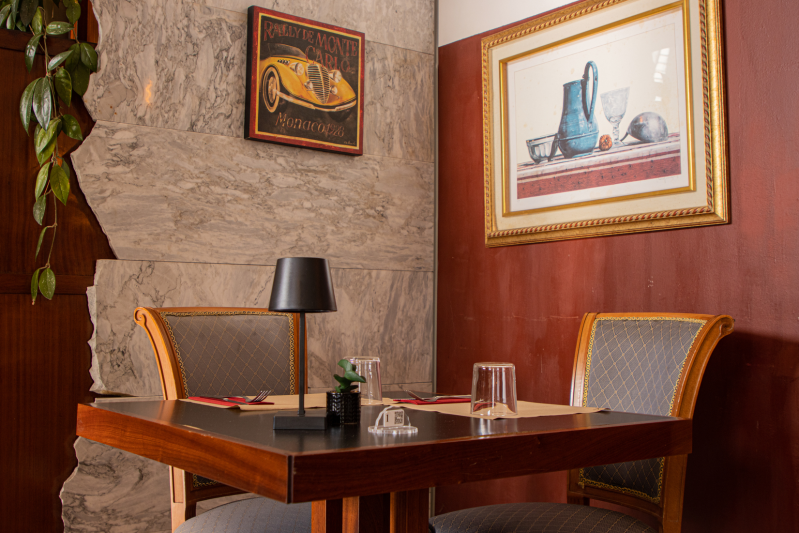 Tavolo da ristorante apparecchiato per due, quadri incorniciati su pareti in mattoni e rosse, una lampada e sedie.