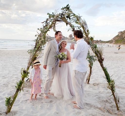 A bride and groom are standing under an arch on the beach