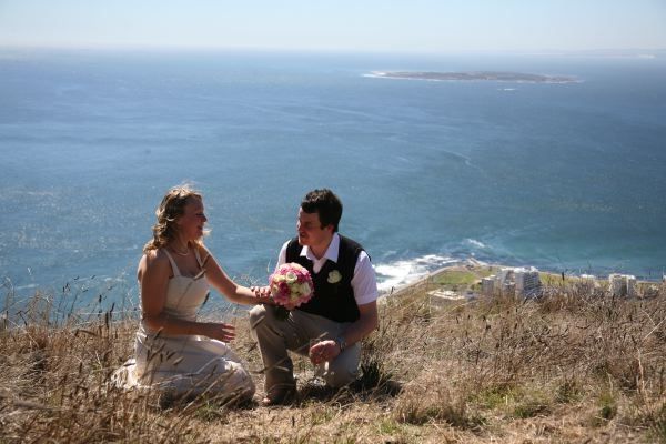 A bride and groom are sitting on top of a hill overlooking the ocean.