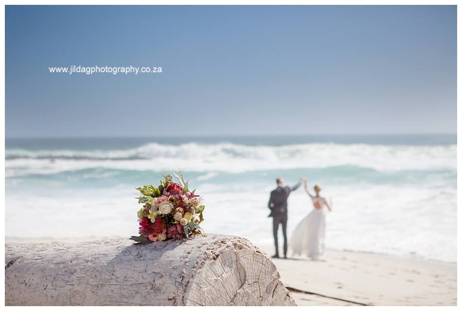 A bride and groom are standing on a beach with a bouquet of flowers in the foreground.