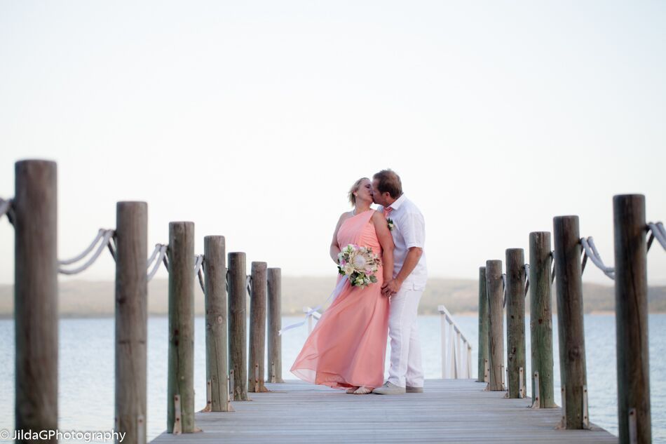 A bride and groom are kissing on a dock overlooking the ocean.
