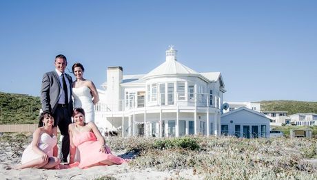 A group of people posing for a picture on a beach in front of a large white house.