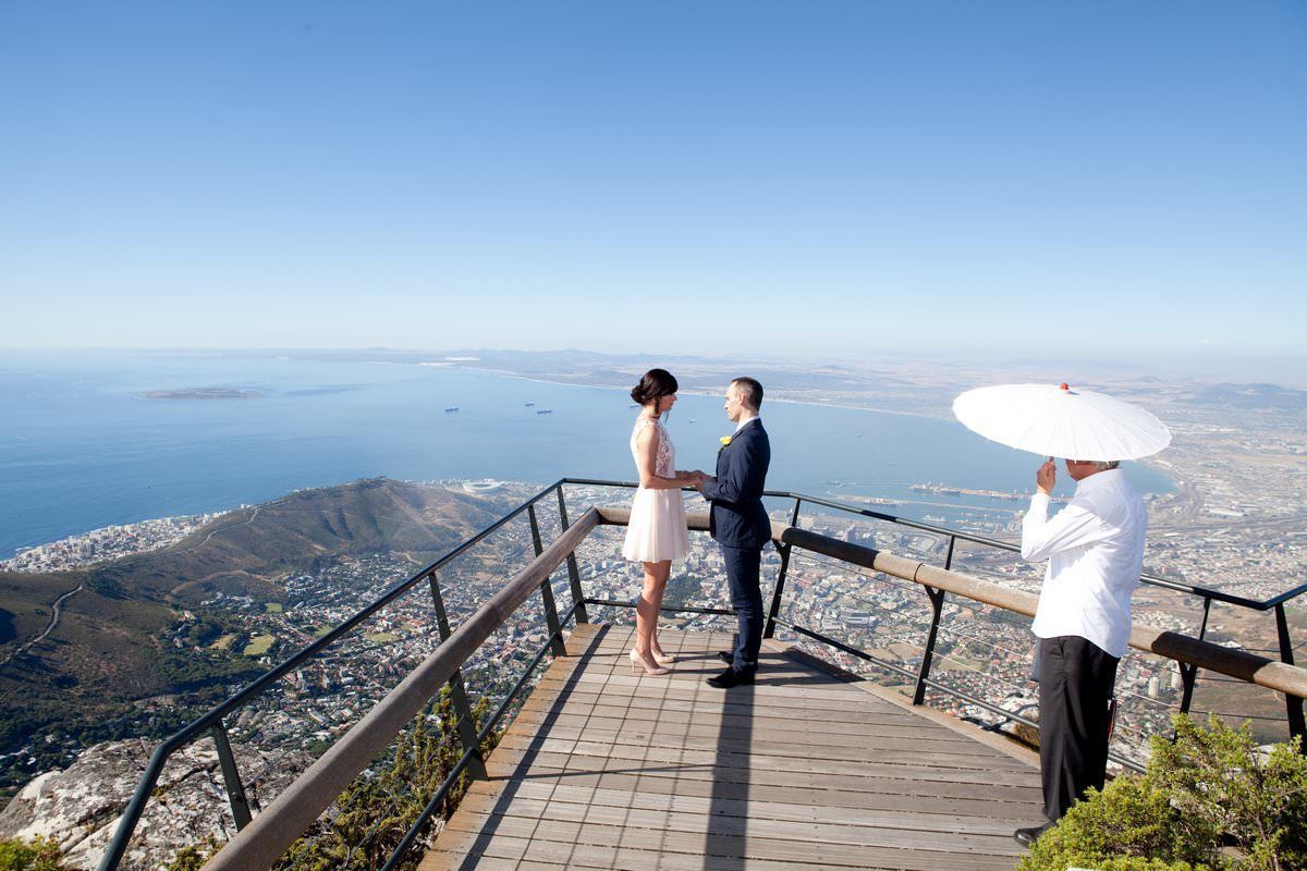 A man is taking a picture of a bride and groom on top of a mountain.