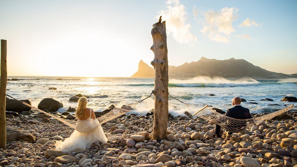 A bride and groom are standing on a rocky beach at sunset.