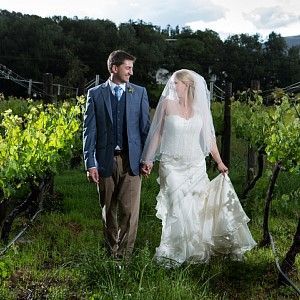 A bride and groom are walking through a vineyard holding hands.