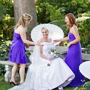 A bride and her bridesmaids are sitting on a bench holding umbrellas.