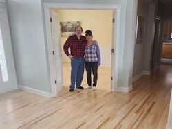 Couple standing in doorway, facing camera. Room with light-colored hardwood floors, framed doorway, and light blue walls.