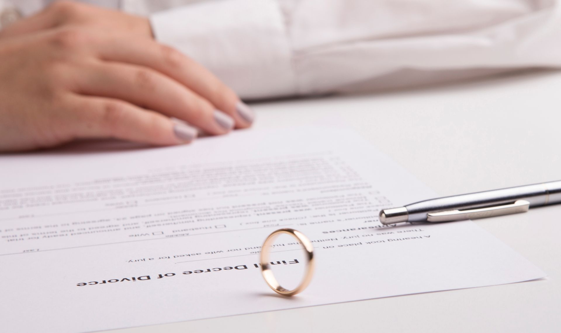 Close-up of a hand near final divorce papers and ring from a divorce attorney's desk. Wedding ring on divorce papers with a hand in the background and a pen.