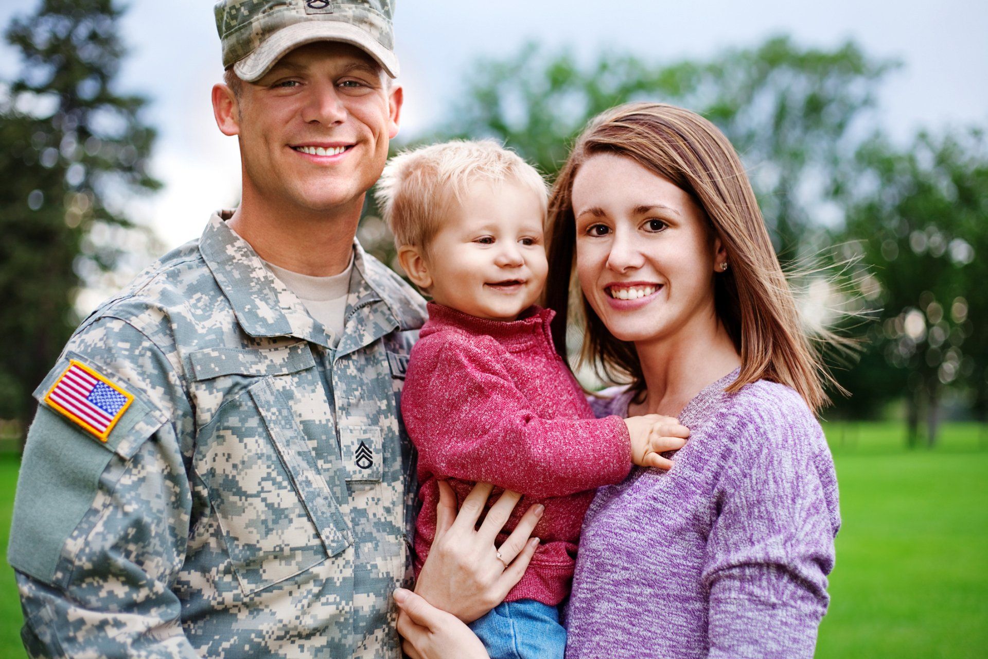 Soldier in uniform smiles with a woman and child on green grass.