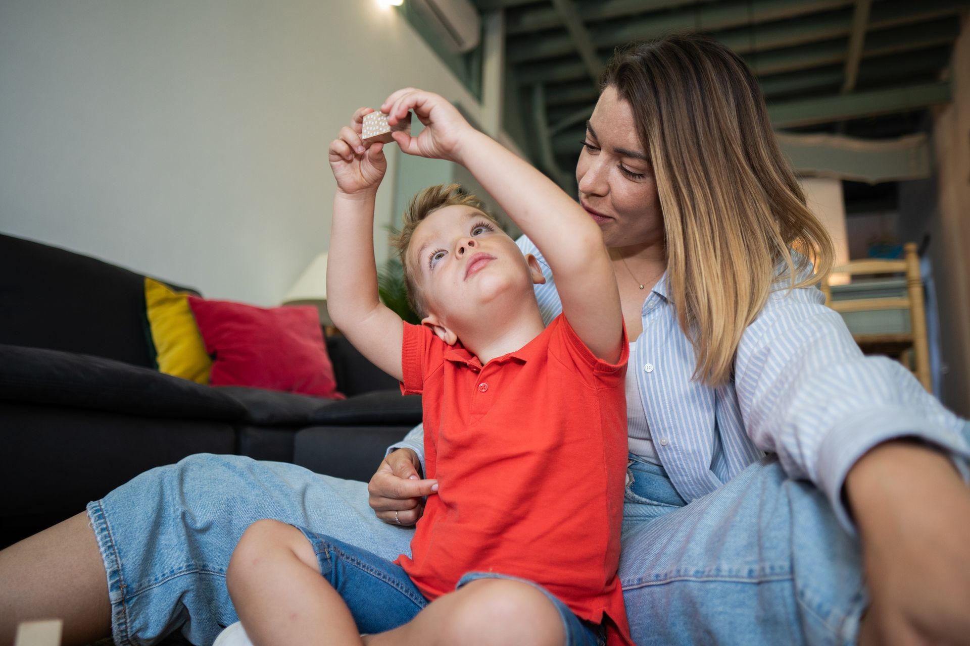 Young boy raising his arms while holding a wooden block with his mother watching him.