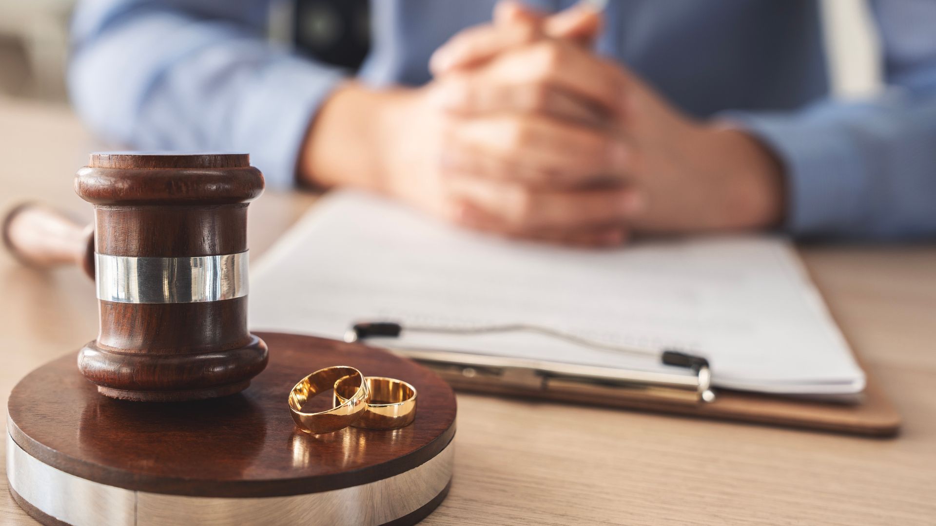 Gavel, wedding rings, and legal documents on a table with a person's hands clasped in the background.