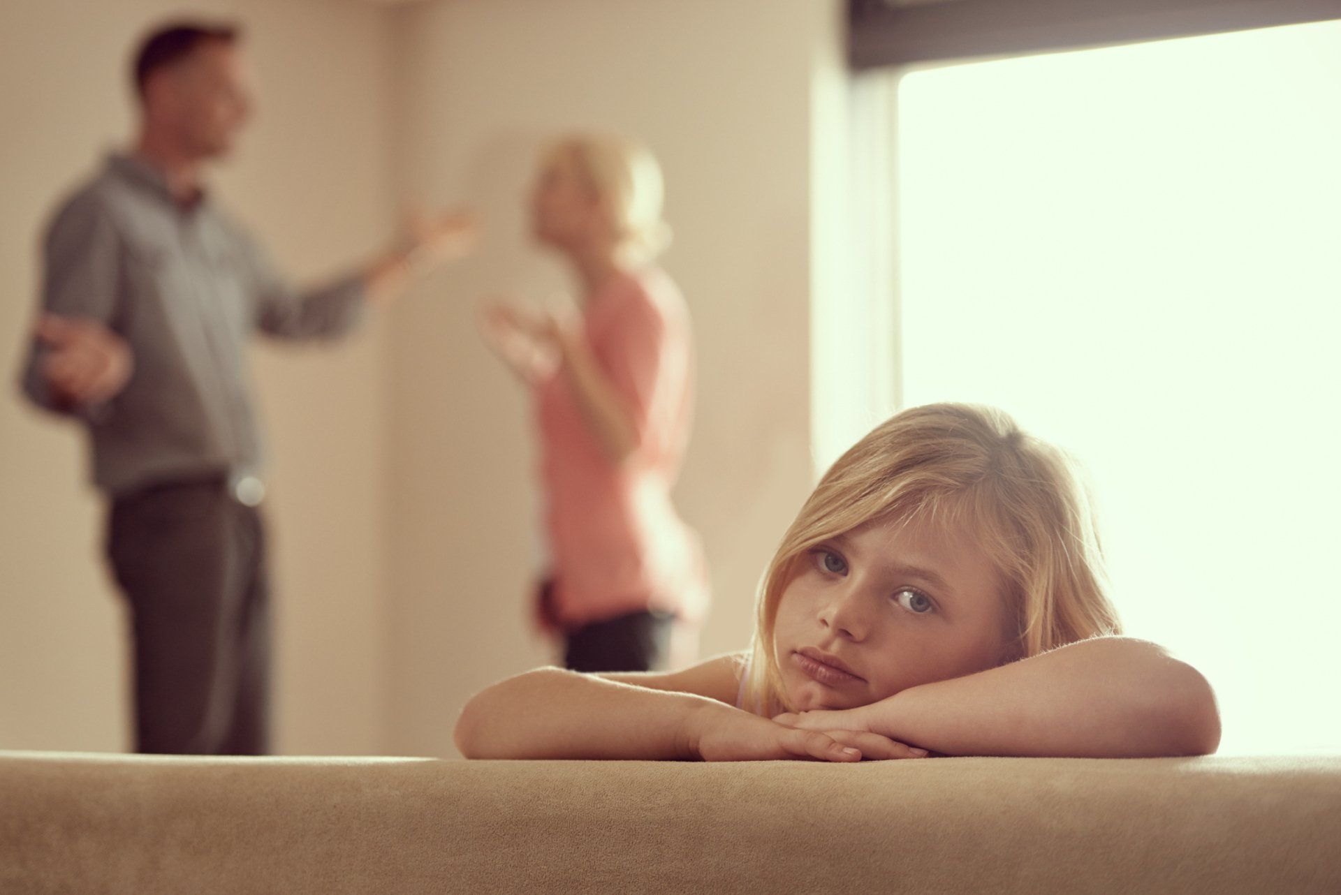 Girl with sad expression in foreground; parents arguing in the blurred background.