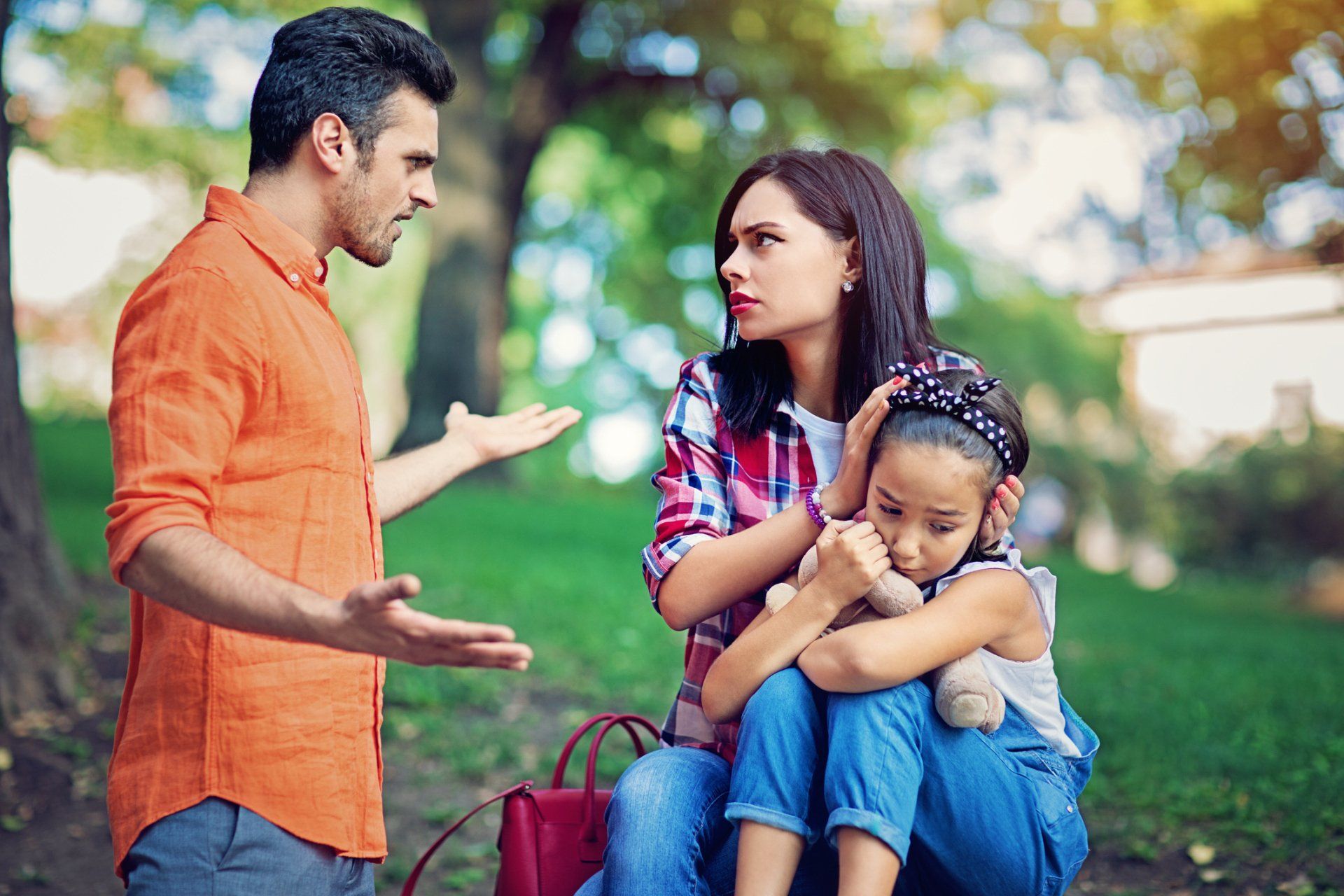 Man and woman arguing outdoors, child covering ears, looking upset.
