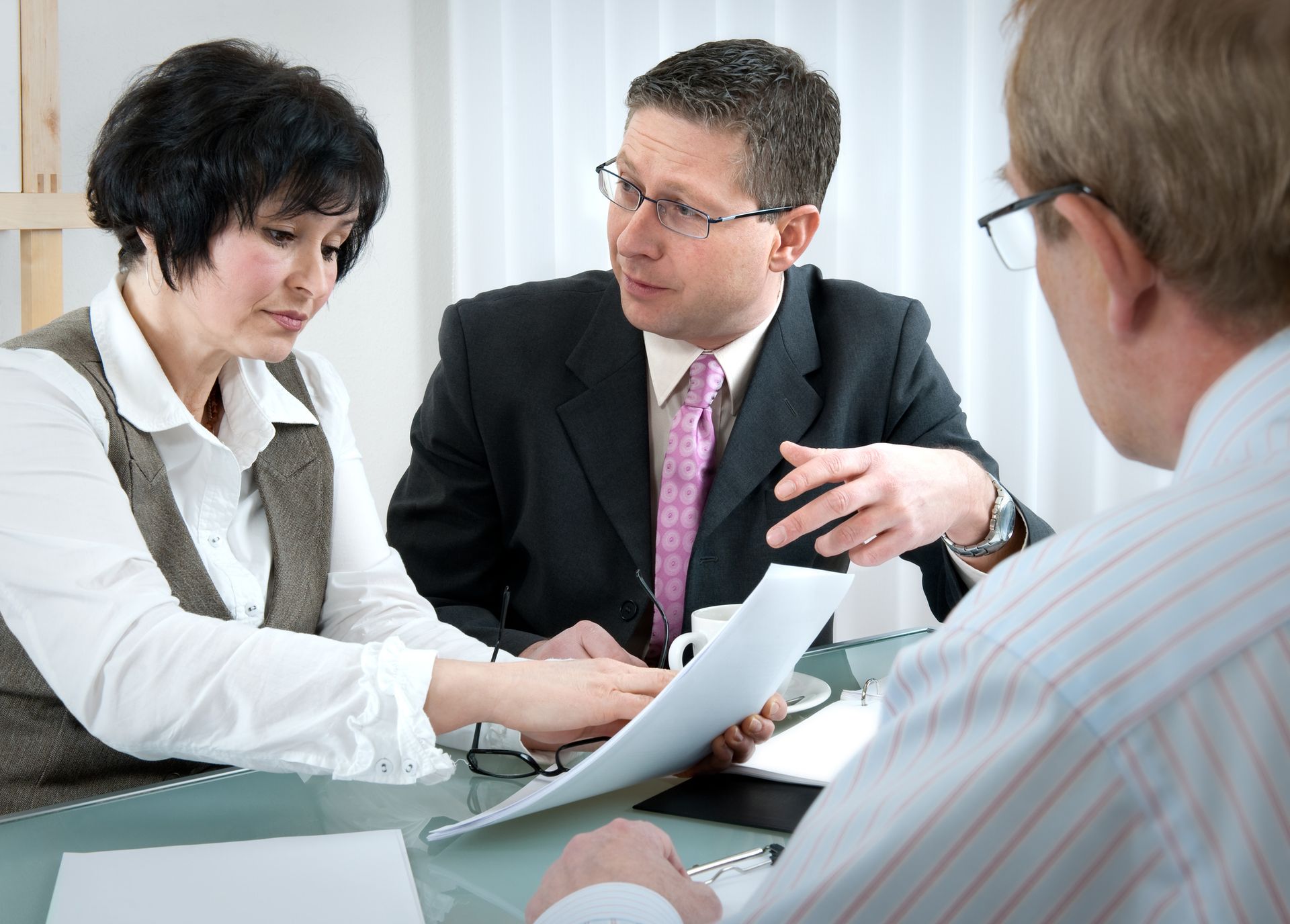 Three people reviewing documents at a table. A man in a suit gestures, explaining paperwork to a woman. Another man is partially visible.