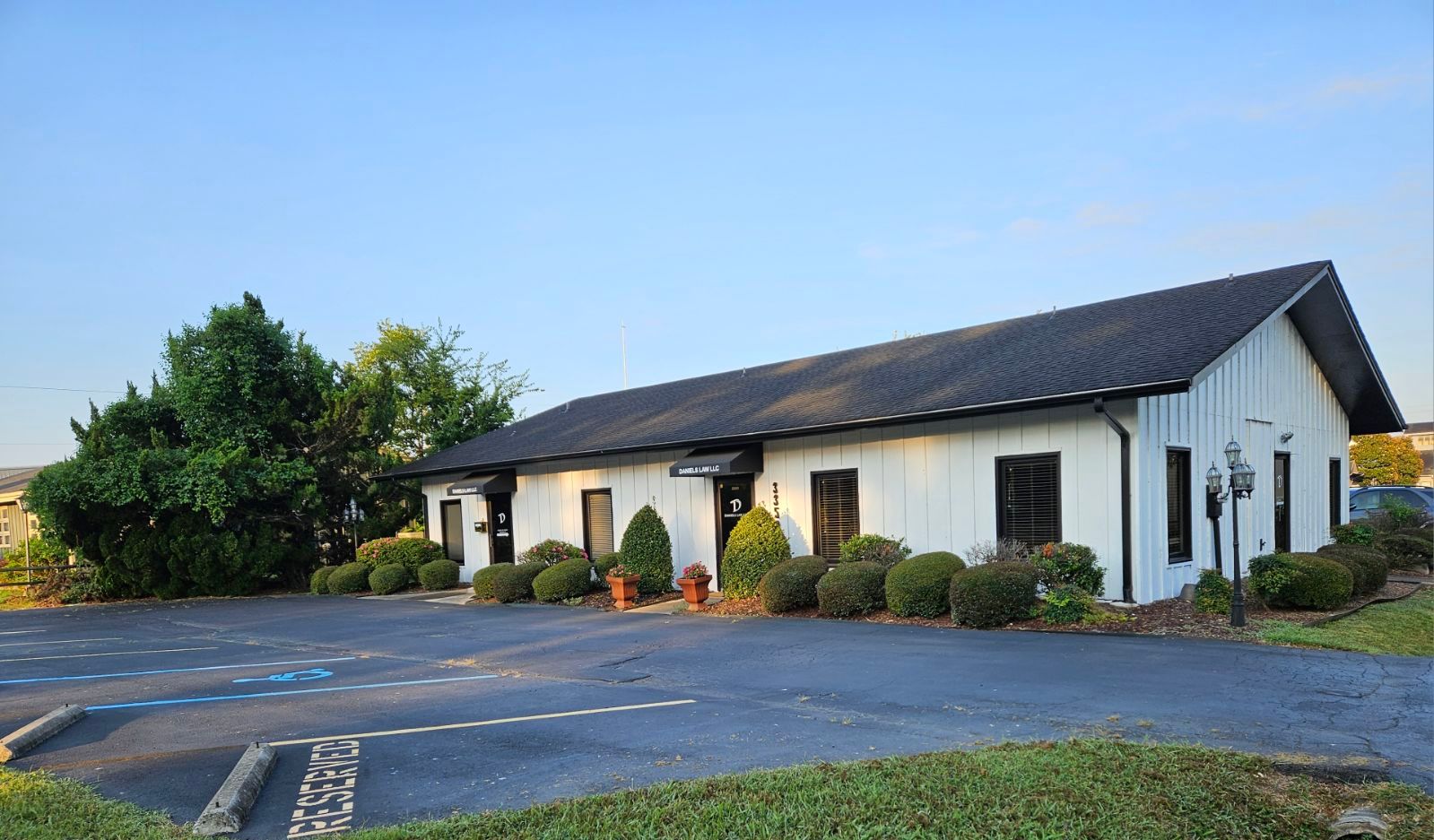 White building with black roof, small windows, bushes, and a parking lot.
