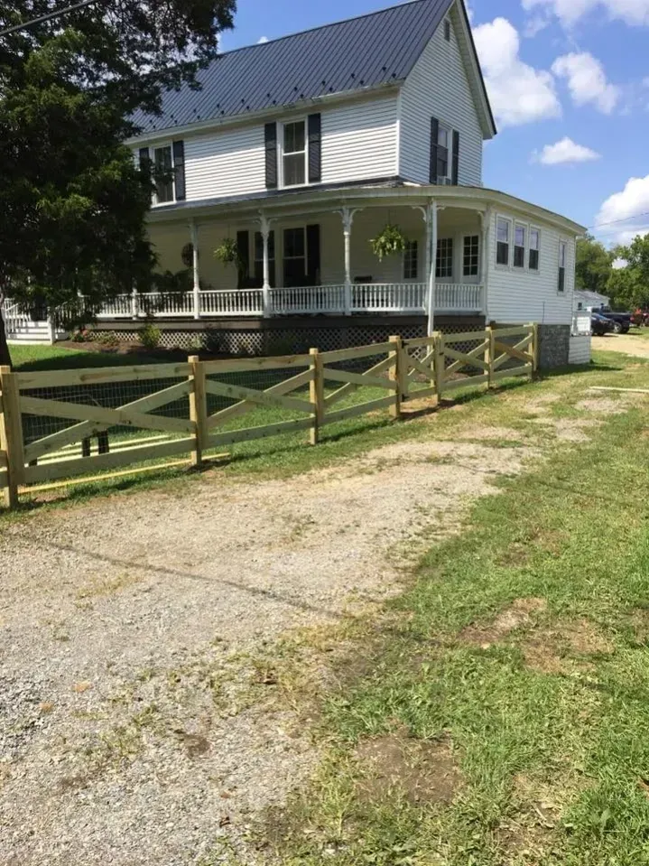 White farmhouse with wraparound porch, wooden fence, and gravel driveway on a sunny day