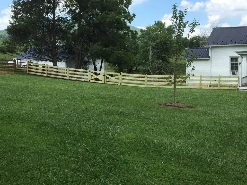 Grassy backyard with a wooden fence, trees, and a white house under a partly cloudy sky