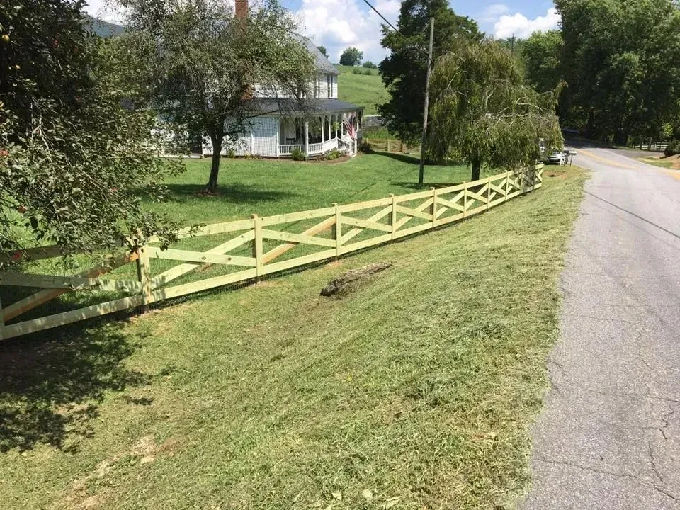 Rural roadside with a white house, trees, and a yellow split-rail fence along the grassy lawn.