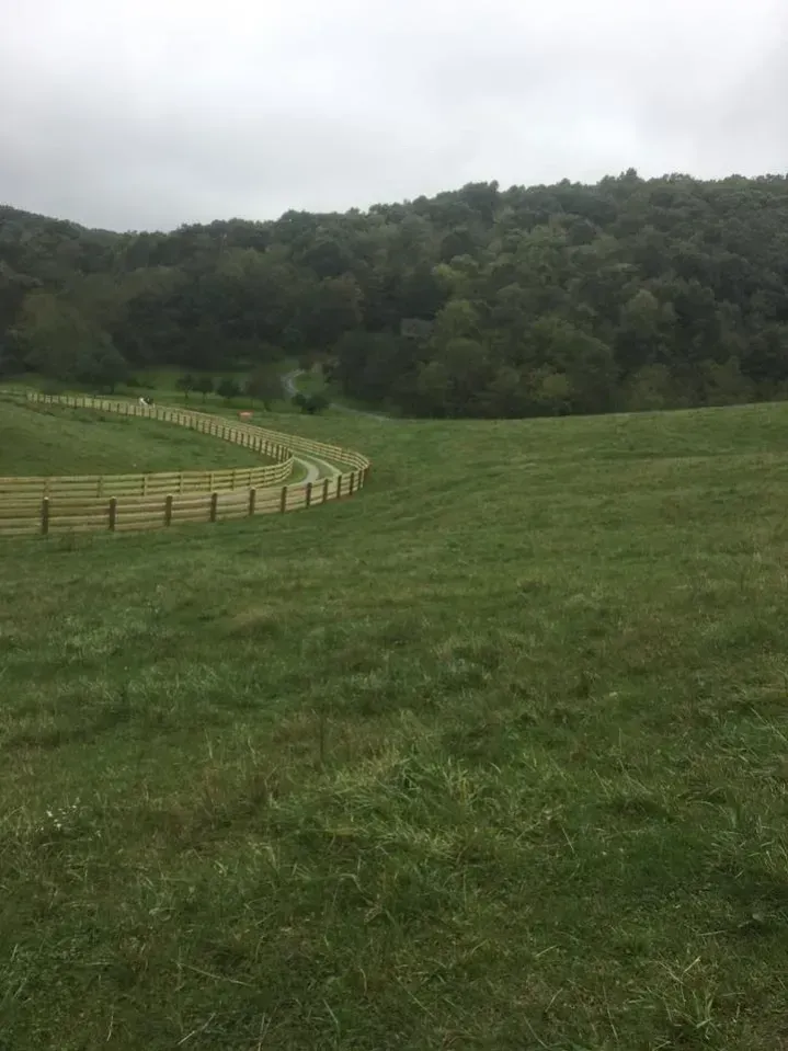 Grassy hillside meadow with a curving wooden fence and tree-covered hills under a cloudy sky