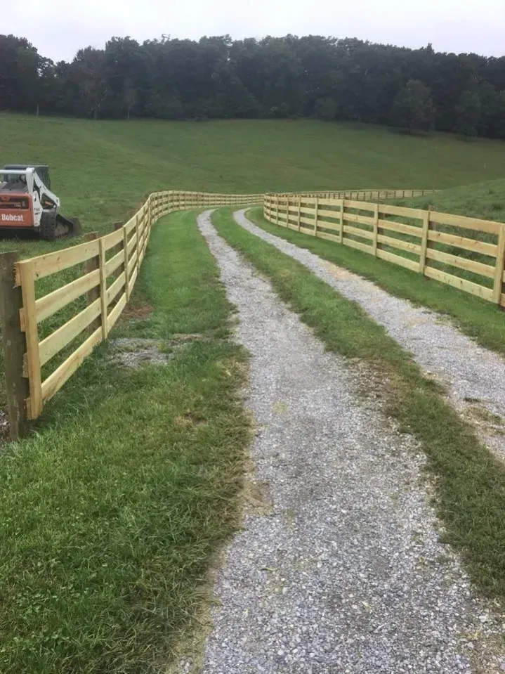 Gravel driveway between wooden fences leading through a grassy field toward trees