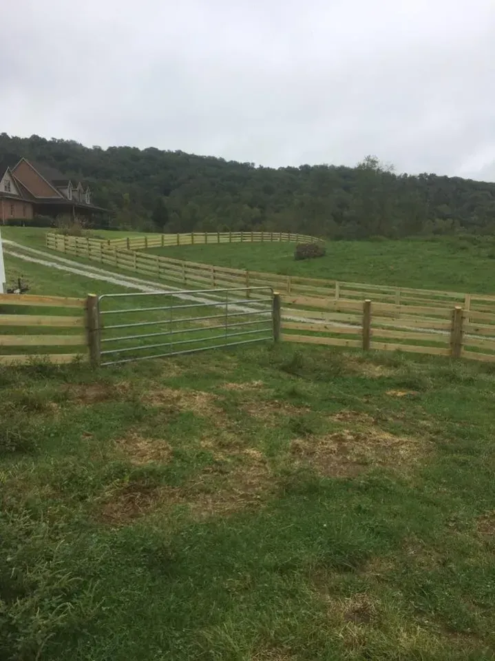 Grassy fenced pasture with a gate and a house near wooded hills under an overcast sky