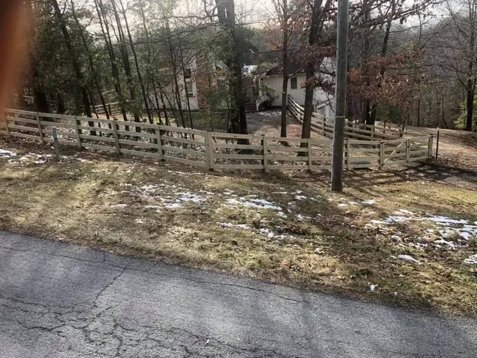Wooded roadside with a wooden fence, patchy snow, and a house in the background
