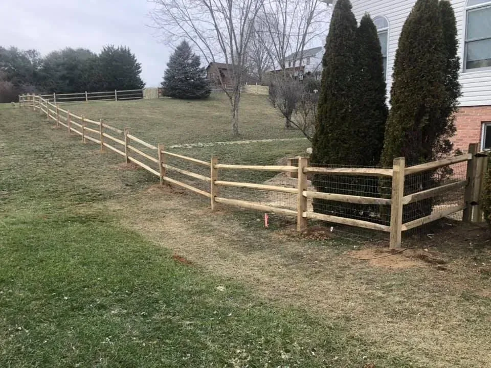 Wooden split-rail fence along a grassy hillside beside evergreen trees and a brick house.
