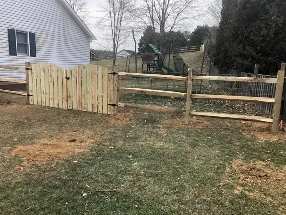 Wooden fence and gate beside a white house in a grassy backyard with trees and a playset.