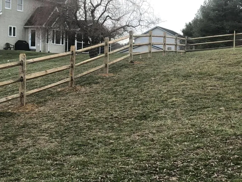 Wooden fence along a grassy hill beside a house and shed on an overcast day
