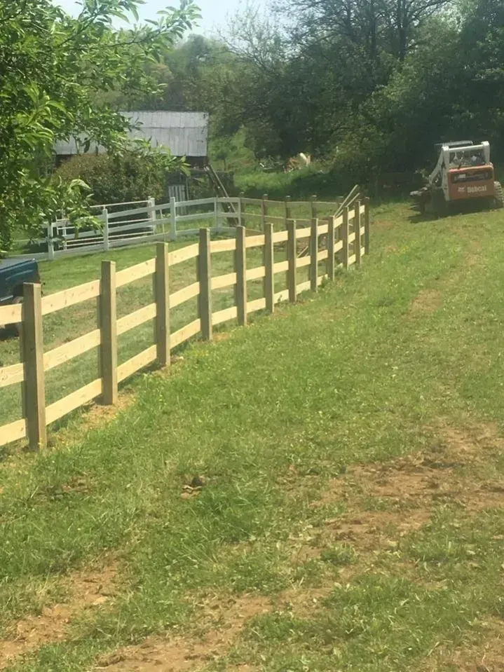 Wooden fence lines a grassy driveway beside trees and a small barn, with a tractor in the distance.