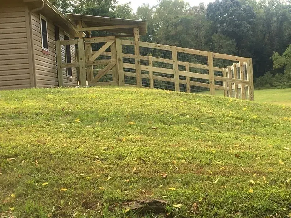 Wooden deck ramp beside a house on a grassy hillside, with a fenced platform and trees in the background