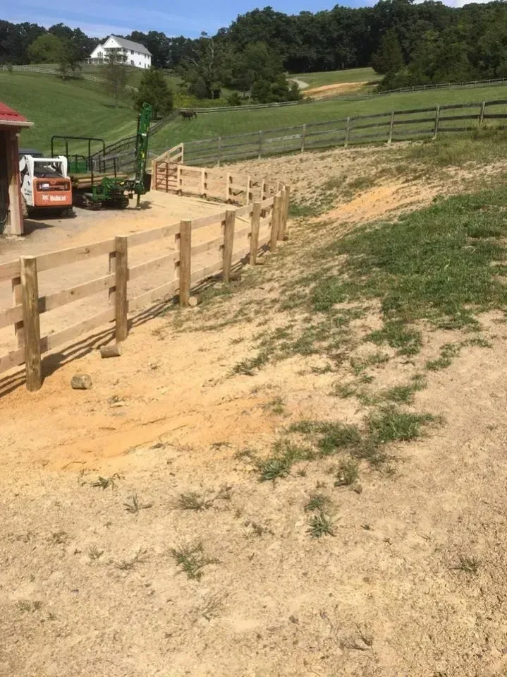 Dirt path beside a wooden fenced slope with a small tractor and grassy hills under a clear sky