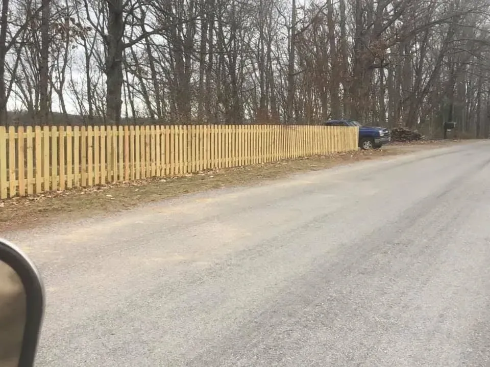Quiet road beside a yellow picket fence, with leafless trees and a parked blue car in the distance