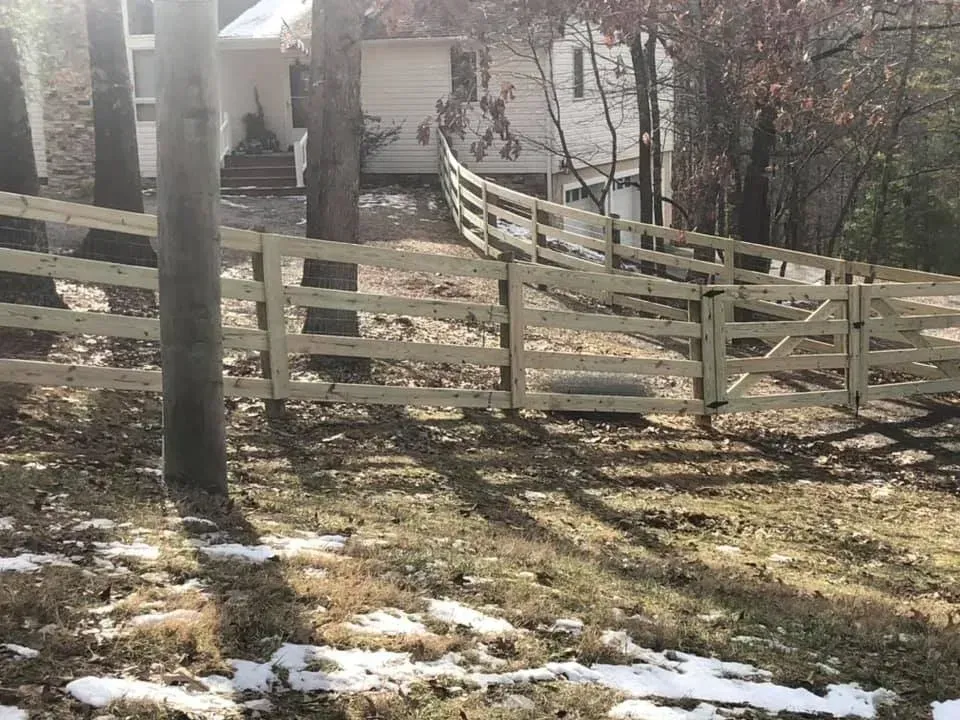 Wooden fence with a gate in a snowy yard, with bare trees and a house in the background.
