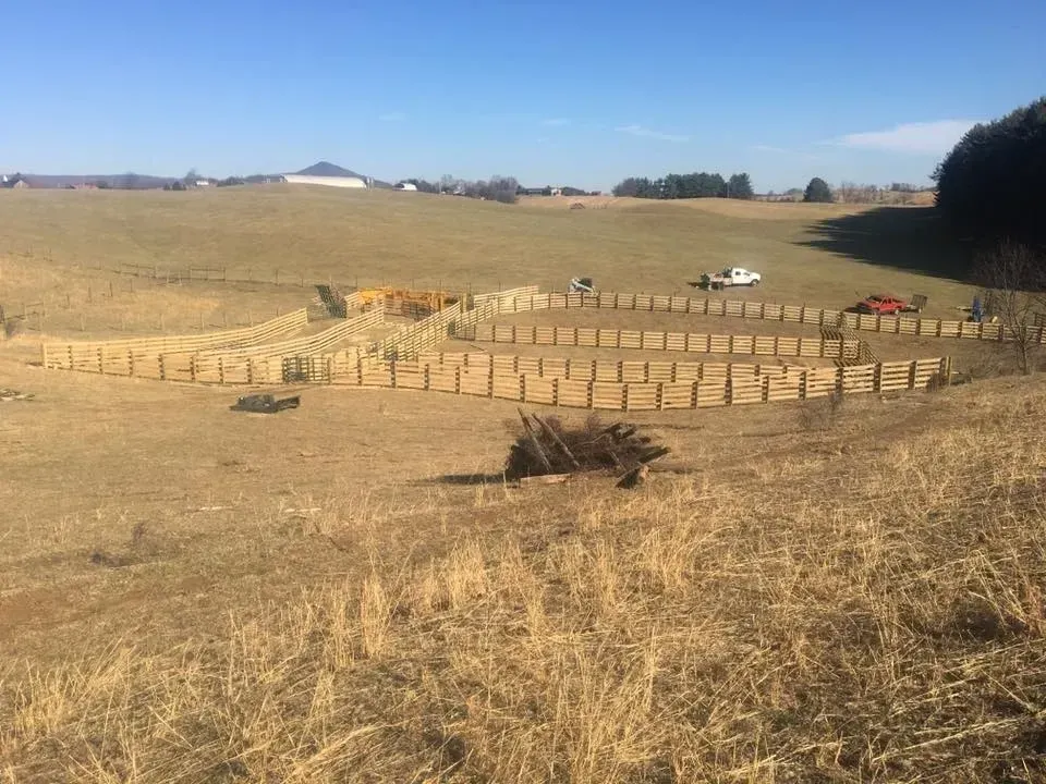 Dry rural field with fenced circular pen, a few parked vehicles, and distant buildings under a blue sky