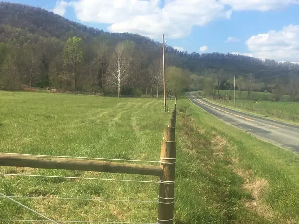 Rural roadside beside a green field, fence posts, hills, and a blue sky.