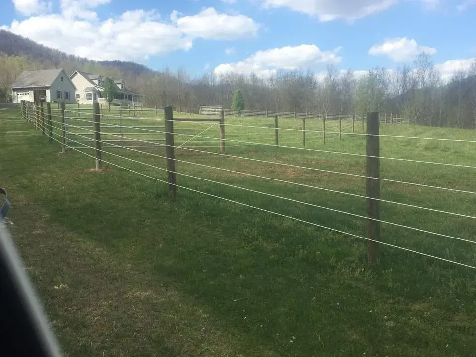 Rural pasture with wooden fence posts and wire fencing under a blue sky, with a house in the distance