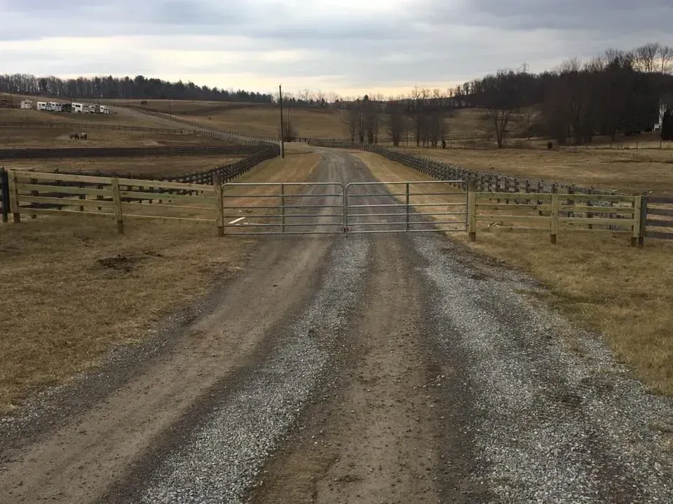 Gravel driveway leading through open fields with wooden fences under a cloudy sky