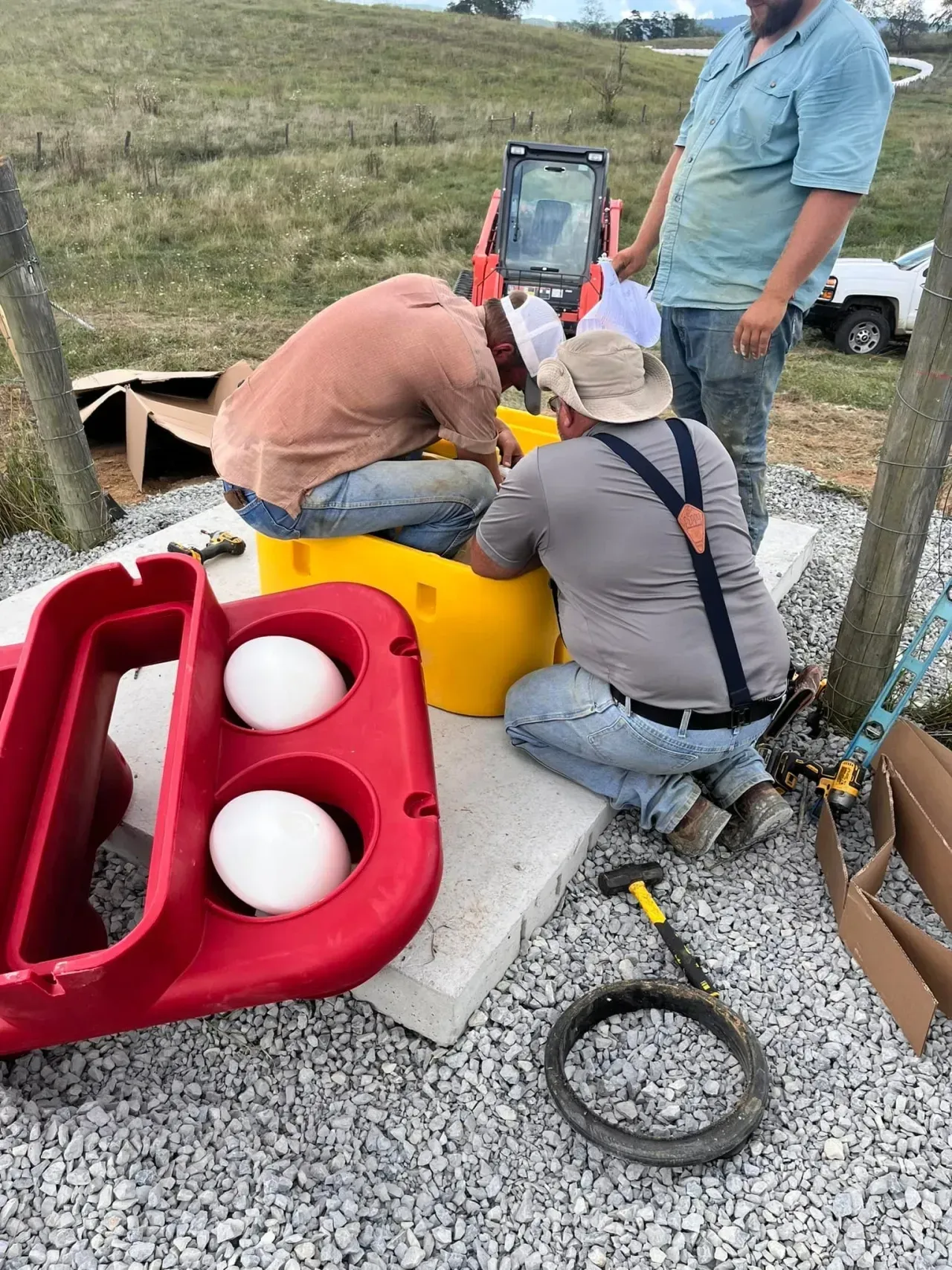 Workers crouch over a yellow utility box outdoors beside a red bin and white tanks.