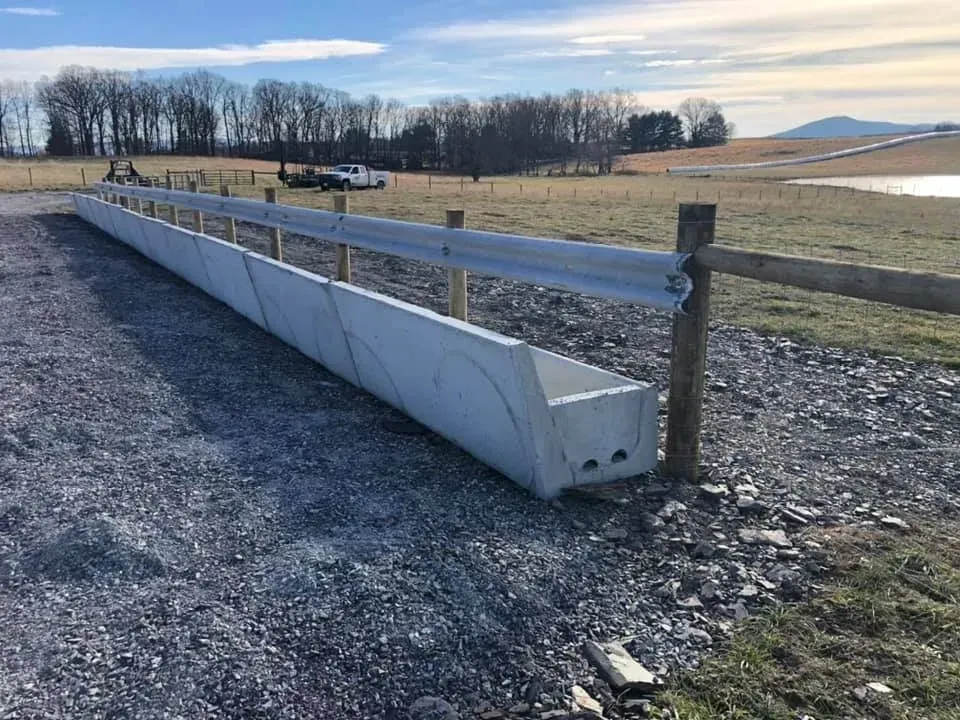 Long concrete barrier along a gravel roadside beside a fence in open fields under a blue sky