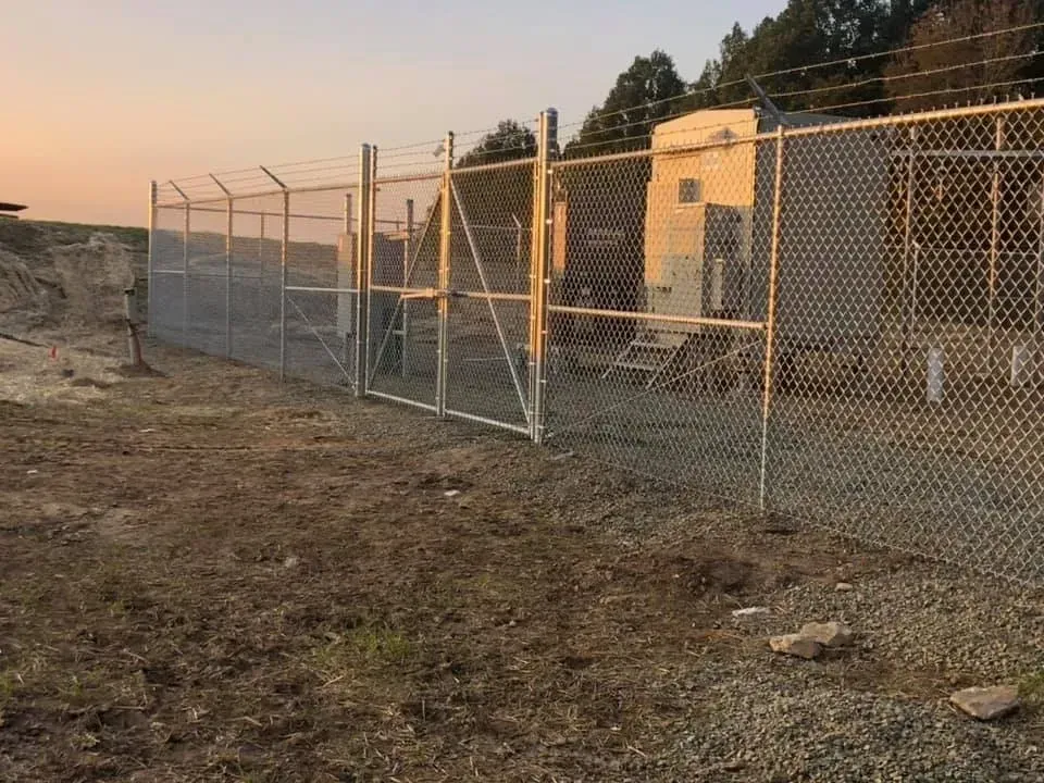 Chain-link fence beside a dirt path at sunset, with trees and a small building in the background.