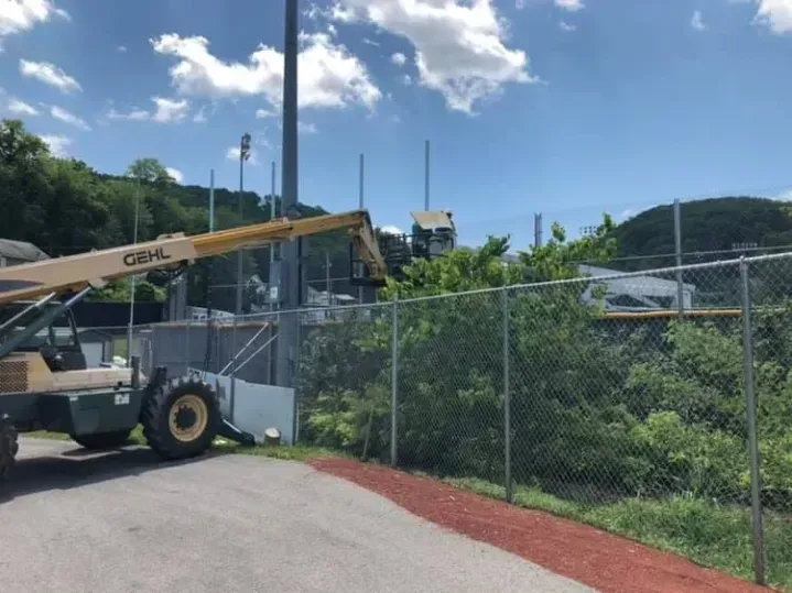 Construction lift beside chain-link fence under blue sky, with green vegetation and road in foreground