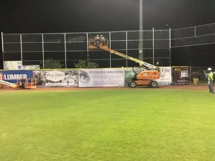 Nighttime sports field with a cherry picker beside the outfield fence and banners along the wall