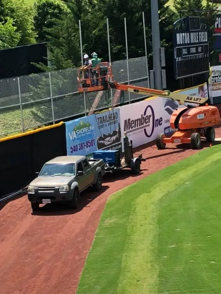 Orange cherry picker lifts worker over baseball field wall beside trucks and ads on the warning track