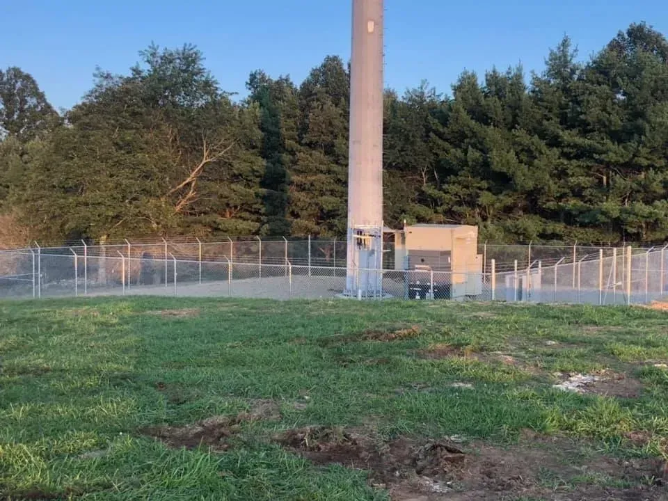 Grassy field with a fenced utility tower and trees in the background