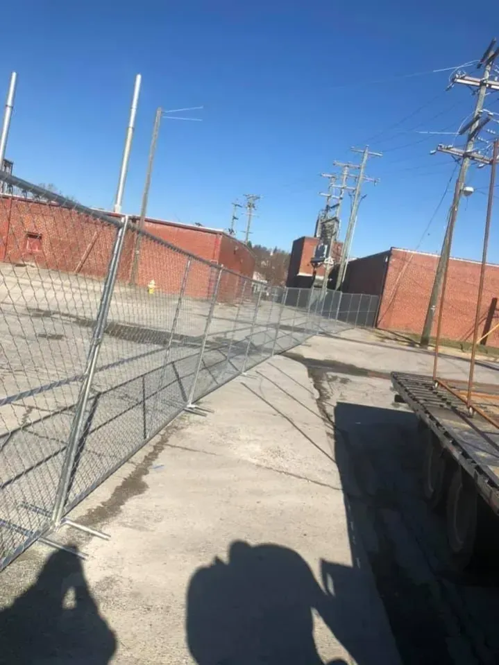 Chain-link fence along a concrete walkway beside red brick buildings under a clear blue sky.