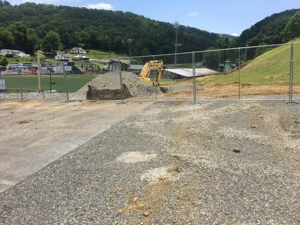 Construction site with excavator, gravel foreground, and fenced grassy hillside under clear sky