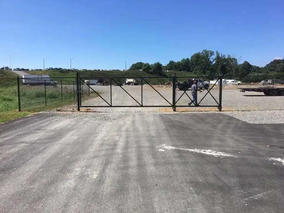 Chain-link gate across a gravel lot under a clear blue sky with a few parked cars beyond.