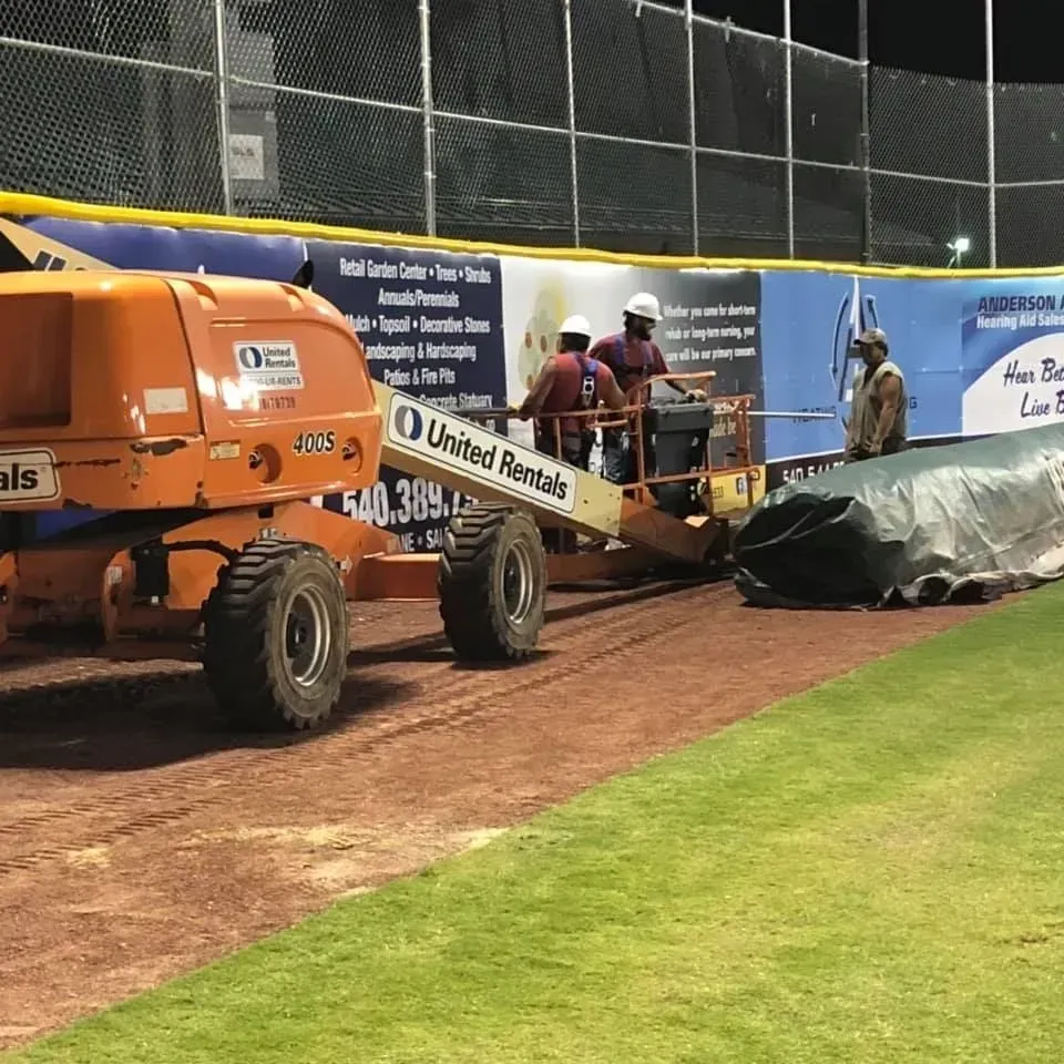 Orange lift on baseball field beside tarp-covered mound and workers near dugout at night