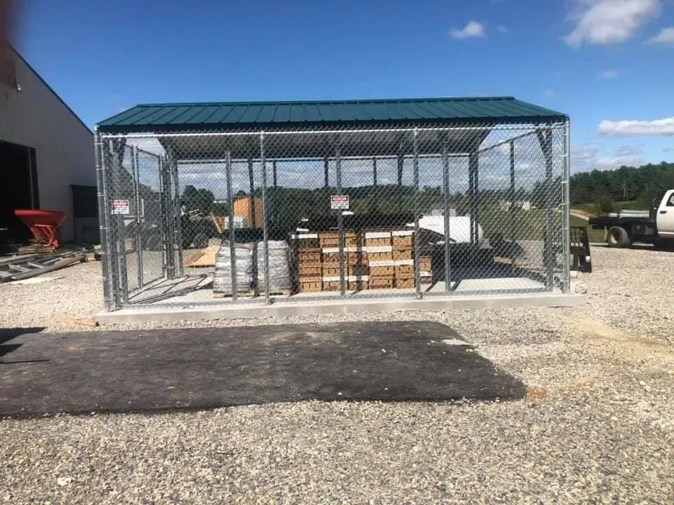 Chain-link storage enclosure with a green roof, pallets inside, on a gravel lot beside a building.